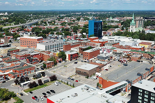 Vue aérienne du centre-ville de Trois-Rivières comportant des immeubles commerciaux et résidentiels, des rues quadrillées et des zones de stationnement, sous un ciel partiellement nuageux.